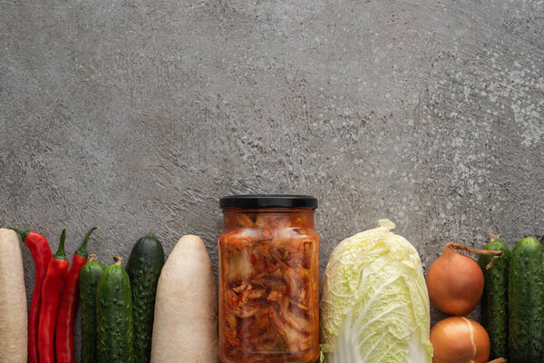 top view of chili peppers, cucumbers, daikon radish, chinese cabbage, onions and kimchi jar on grey concrete background