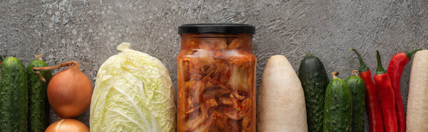 panoramic shot of chili peppers, cucumbers, daikon radish, chinese cabbage, onions and kimchi jar on grey concrete background