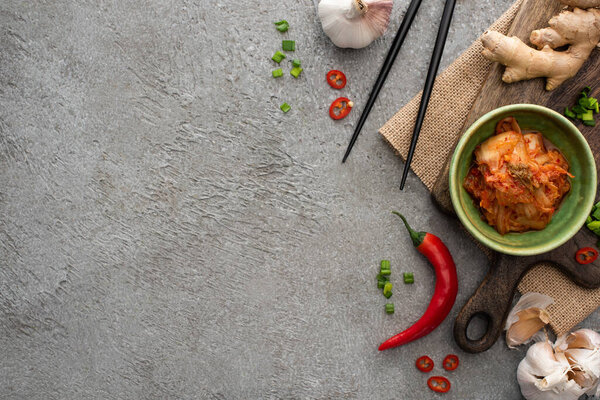 top view of bowl with kimchi on cutting board near chopsticks, ginger, chili pepper and garlic on concrete surface
