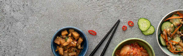 panoramic shot of bowls with delicious kimchi near sliced cucumber and chopsticks on concrete surface