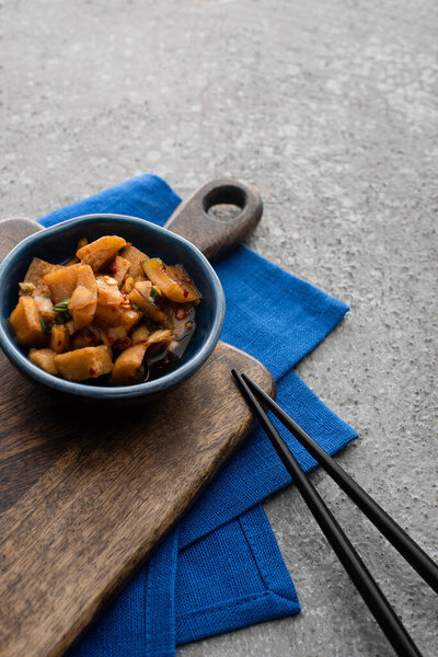 bowl with deicious kimchi on wooden cutting board and blue cloth near chopsticks on concrete surface