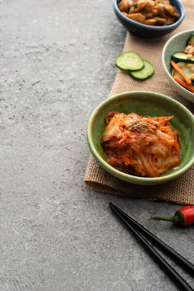 bowls of tasty kimchi on sackcloth near chopsticks on concrete surface