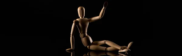 Wooden doll in sitting position with outstretched hand and crossed legs on black background, panoramic shot
