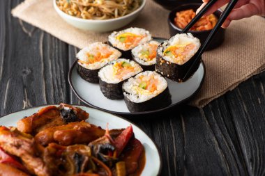 cropped view of woman holding chopsticks with rice roll near korean side dishes  clipart