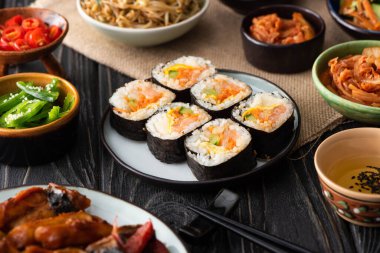 selective focus of gimbap with salmon near chopsticks and korean side dishes  clipart