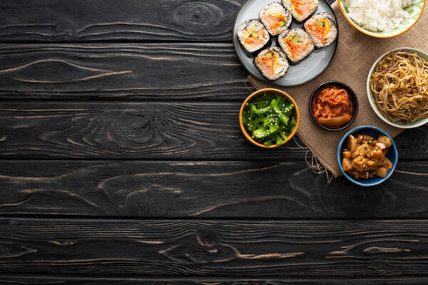 top view of bowls with tasty korean side dishes near gimbap on wooden surface 