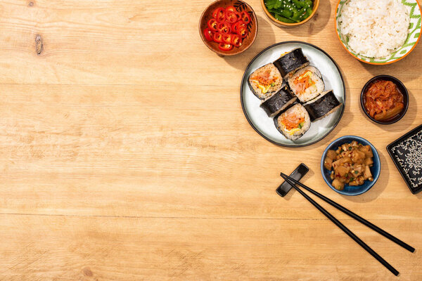 top view of bowls with tasty korean side dishes near chopsticks and gimbap on wooden surface 