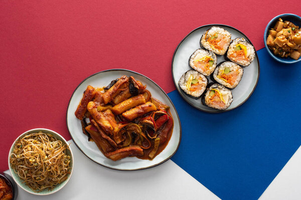 top view of fresh tteokbokki  and gimbap near korean side dishes on white, blue and crimson 