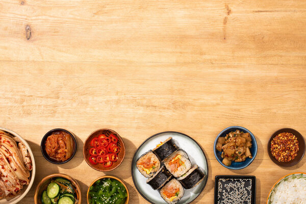 top view of bowls with korean side dishes near korean gimbap on wooden surface 