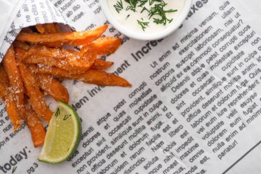 top view of crispy french fries, sliced lime and garlic sauce with dill on newspaper  clipart