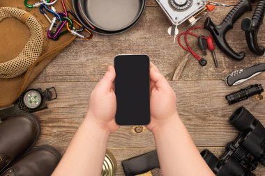 Ahşap masadaki yürüyüş ekipmanlarının üzerinde boş ekranlı akıllı telefonu tutan adamın kısmi görüntüsü