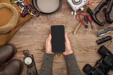 Ahşap masadaki yürüyüş malzemelerinin üzerinde boş ekranlı akıllı telefon tutan kadının kısmi görüntüsü