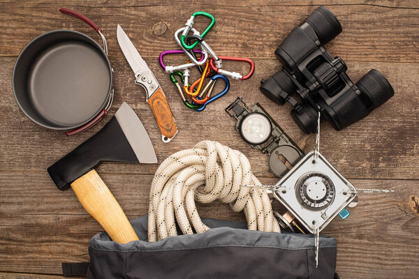 top view of hiking rope, axe, binoculars, knife, carabiners, compass, iron mug and gas burner on wooden table