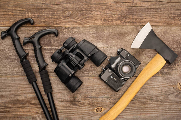 top view of trekking poles, binoculars, photo camera and axe on wooden surface