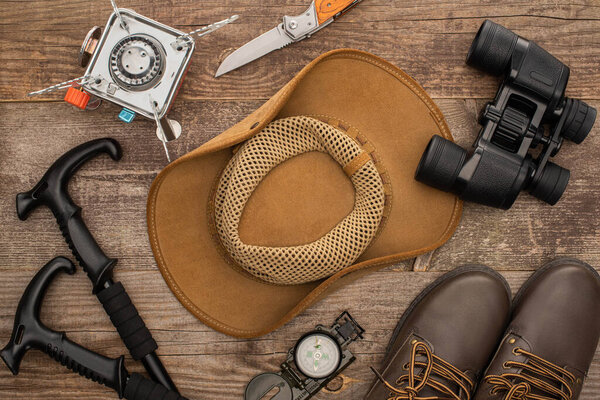 top view of hat, boots, hiking poles, binoculars, jackknife and gas-burner on wooden surface