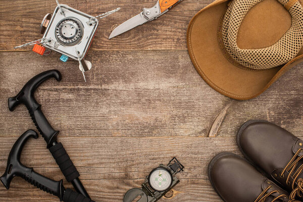 top view of trekking poles, gas burner, jackknife, hat, boots and compass on wooden table