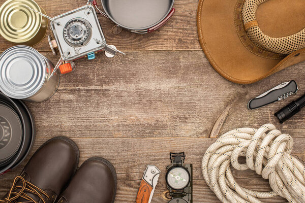 top view of gas burner, metal dishes, tin cans, boots, hat and hiking equipment on wooden surface