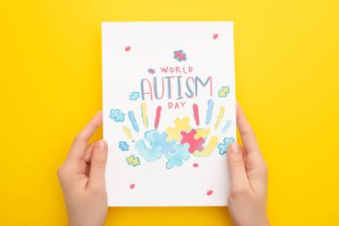 cropped view of woman holding card with puzzle and hand prints for World Autism Day on yellow clipart