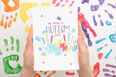 cropped view of woman holding card with World Autism Day lettering and puzzle on white with hand prints clipart