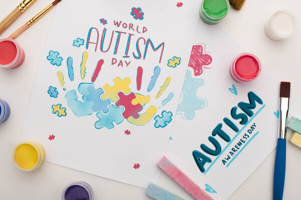 верхний вид открыток ко Всемирному дню аутизма с надписью и покраской головоломок и отпечатков рук на белом с кистями, мелом и красками
