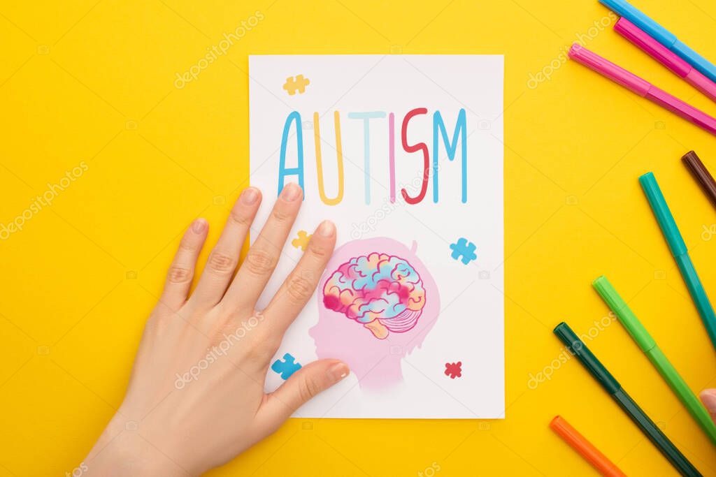top view of person holding paper sheet with autism lettering and drawing of child near felt pens on yellow