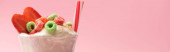 Egy pohár tejturmix jégkrémmel, eper felével, cukorkával és szívószállal rózsaszín alapon, panorámás felvétel