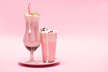 Disposable cup and glass of strawberry milkshakes on plate on pink background clipart