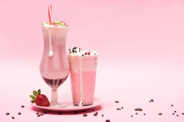 Disposable cup and glass of milkshakes with strawberry on plate and coffee grains on pink background clipart