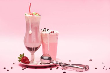 Disposable cup and glass of milkshakes, strawberry and scoop on plate with coffee grains on pink  clipart