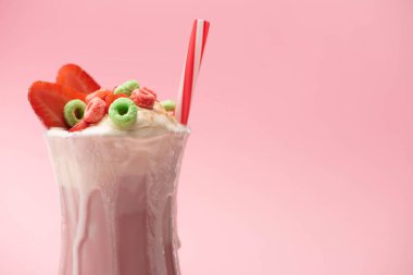 Bir bardak dondurmalı milkshake, renkli şekerler, çilek yarıları ve pembe arka planda içme tüpü.