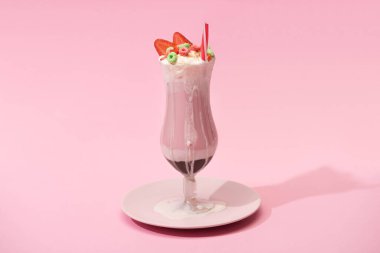 Glass of milkshake with strawberry halves, candies and drinking straw on plate on pink background clipart
