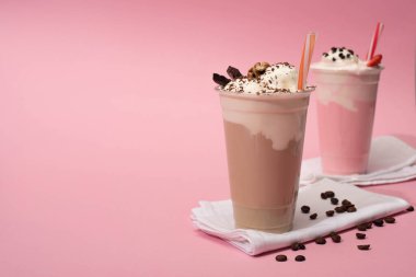 Selective focus of disposable cups of chocolate and strawberry milkshakes with coffee grains on napkins on pink background clipart