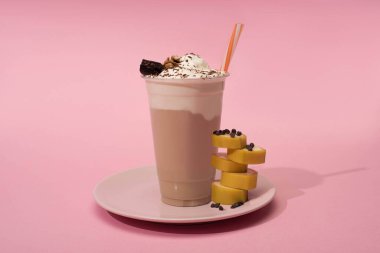 Pembe arka planda tek kullanımlık milkshake, pipet, muz ve çikolata parçacıkları.