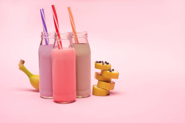 Bottles of berry, strawberry and chocolate milkshakes with drinking straws and cut banana with chocolate chips on pink background