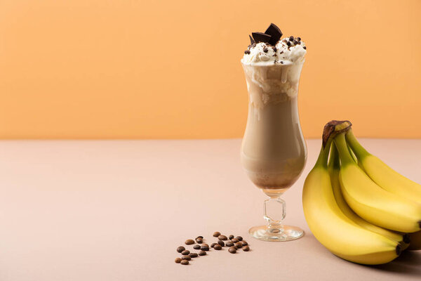 Glass of milkshake with chocolate morsels near bananas and coffee grains on beige and orange