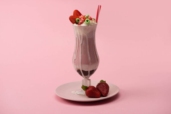 Glass of milkshake with drinking straw, candies and strawberries on plate on pink background