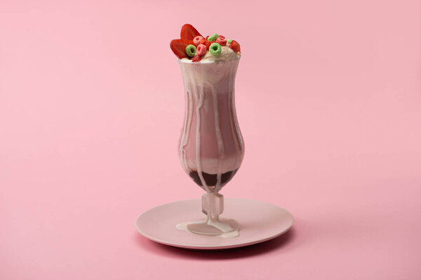 Glass of delicious milkshake with candies and strawberry on plate on pink background 