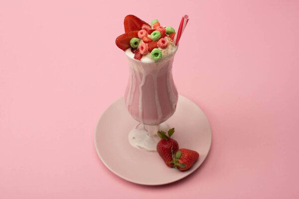 High angle view of glass of milkshake with drinking straw, candies and strawberries on plate on pink background