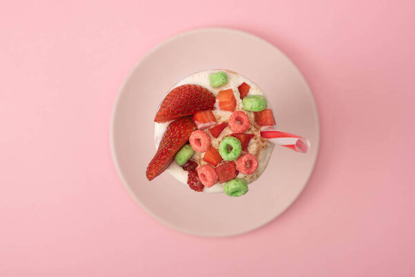 Top view of milkshake with drinking tube, strawberry halves and candies on plate on pink background