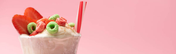 Glass of milkshake with ice cream, strawberry halves, candies and drinking straw on pink background, panoramic shot