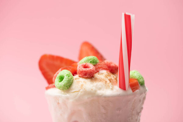 Selective focus of glass of milkshake with ice cream, strawberry halves, candies and drinking straw on pink background