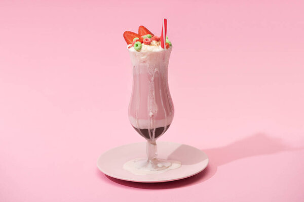 Glass of milkshake with strawberry halves, candies and drinking straw on plate on pink background