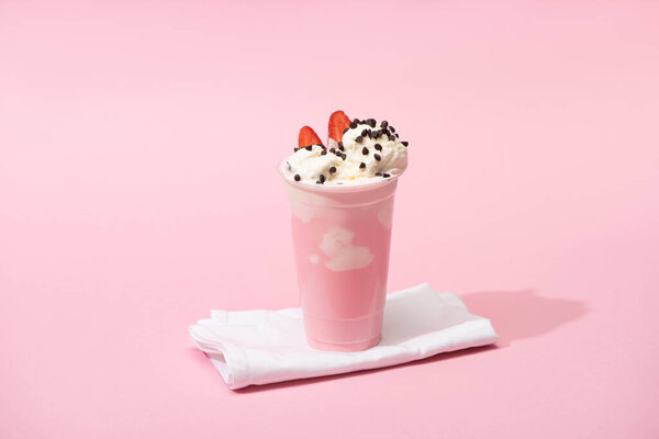 Disposable cup of milkshake with chocolate chips and strawberry halves on napkins on pink background