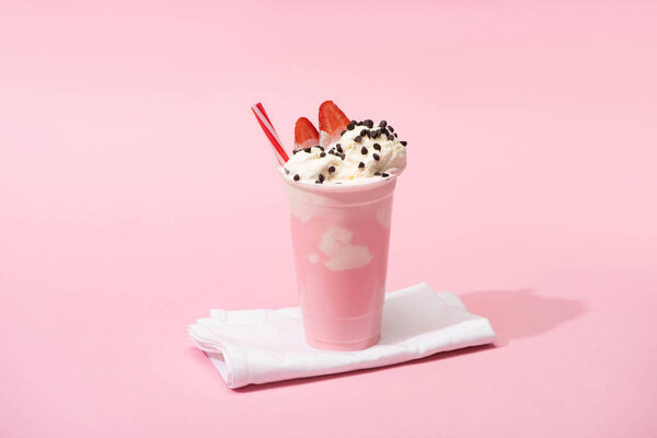 Disposable cup of milkshake with drinking straw, chocolate chips and strawberry halves on napkins on pink 