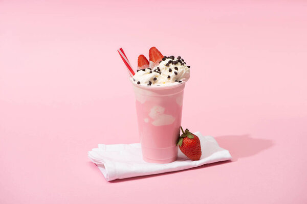 Disposable cup of milkshake with drinking straw, chocolate chips and strawberries on napkins on pink 