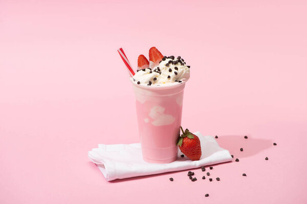 Disposable cup of milkshake with drinking straw, chocolate chips and strawberries on napkins on pink background