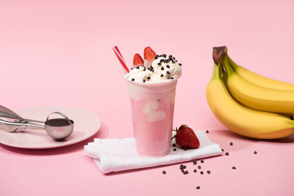 Disposable cup of milkshake with chocolate chips and strawberries on napkins near bananas and plate with scoop on pink 