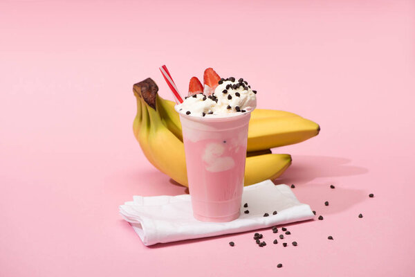 Disposable cup of strawberry milkshake with chocolate chips on napkins near bananas on pink background