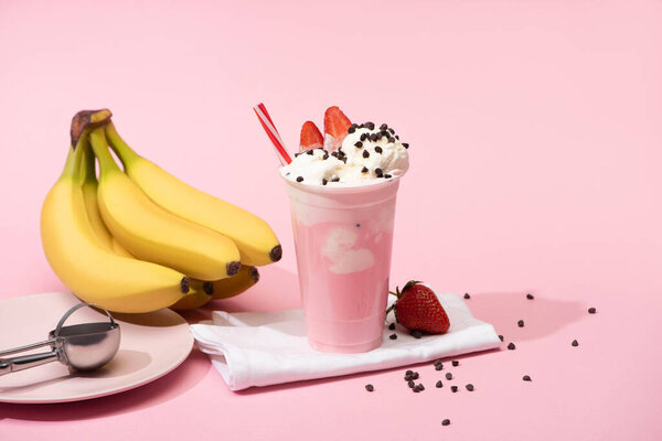 Disposable cup of milkshake with chocolate morsels and strawberries on napkins near bananas and plate with scoop on pink 