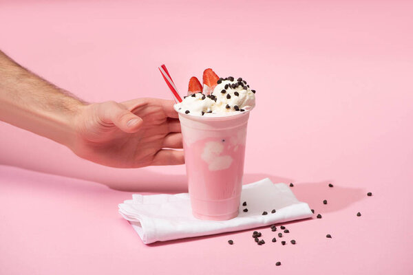 Cropped view of male hand with disposable cup of milkshake with chocolate morsels and strawberry on napkins on pink